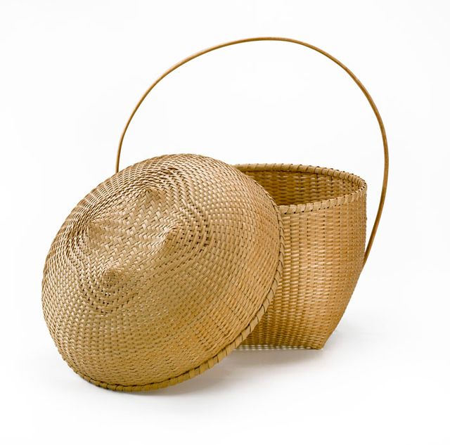 A basket that has a square base that flares our with a circular top and a long thin circular handle. 