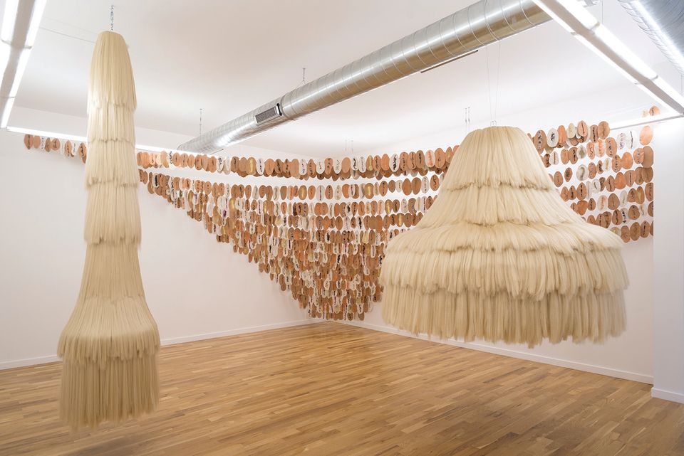 An image of a gallery installation with two objects hanging in the front and a wall hanging in the back.