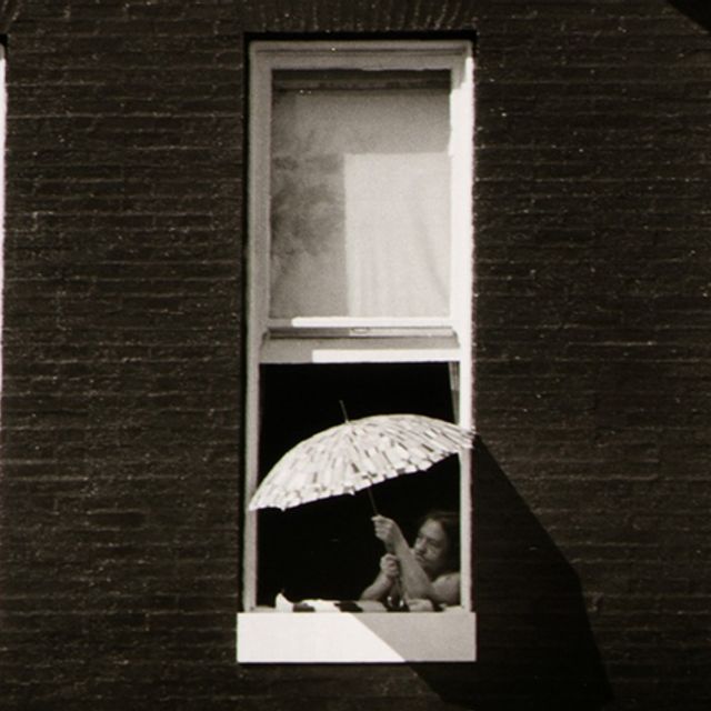 A black and white photograph of a person leaning out of a window, holding an umbrella.