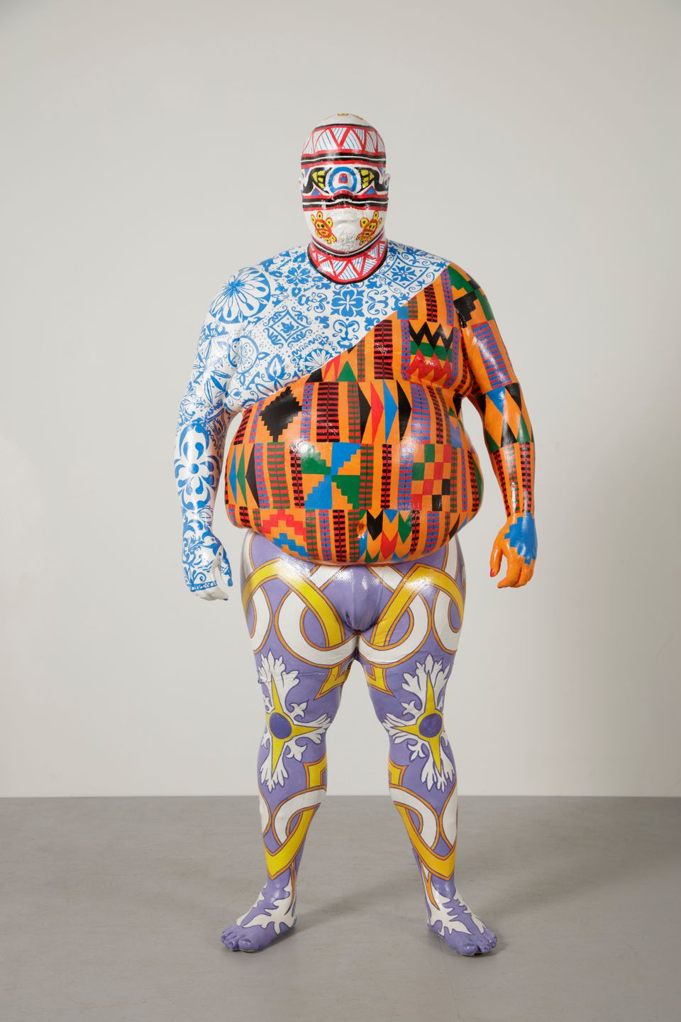 Sculpture of a person completely covered with multiple colorful, intricate patterns standing against a neutral background.