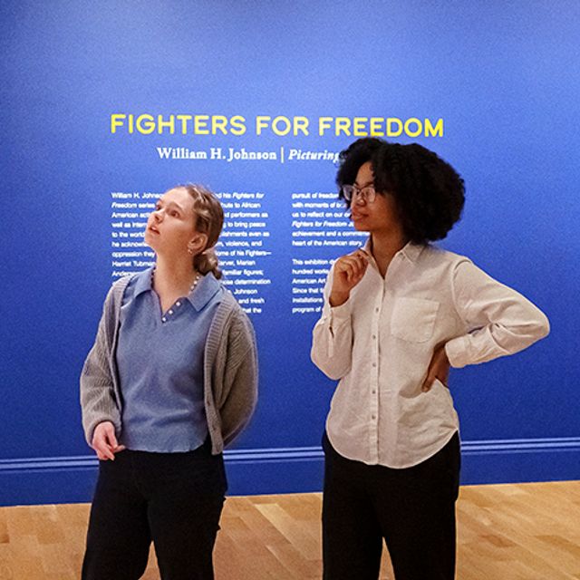 Two visitors look at a timeline on a wall. Behind them is a painting hanging on a blue wall and the words "Fighters for Freedom" with text underneath.