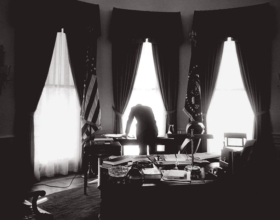 Kennedy bending over in silhouette against the Oval Office windows