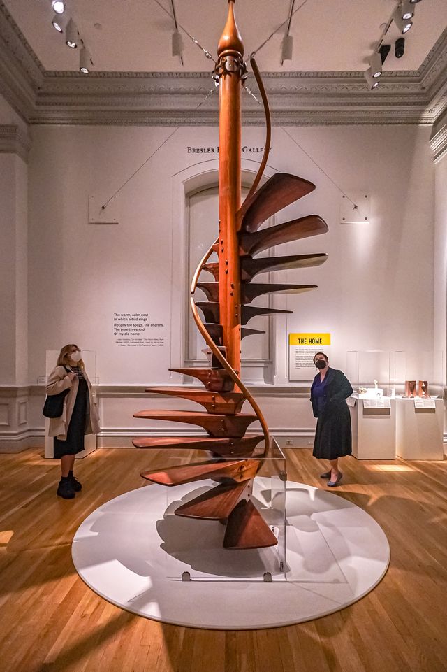 A large sculpture in the shape of a spiral staircase stands at the center of a gallery. Two onlookers in the background look up at it.