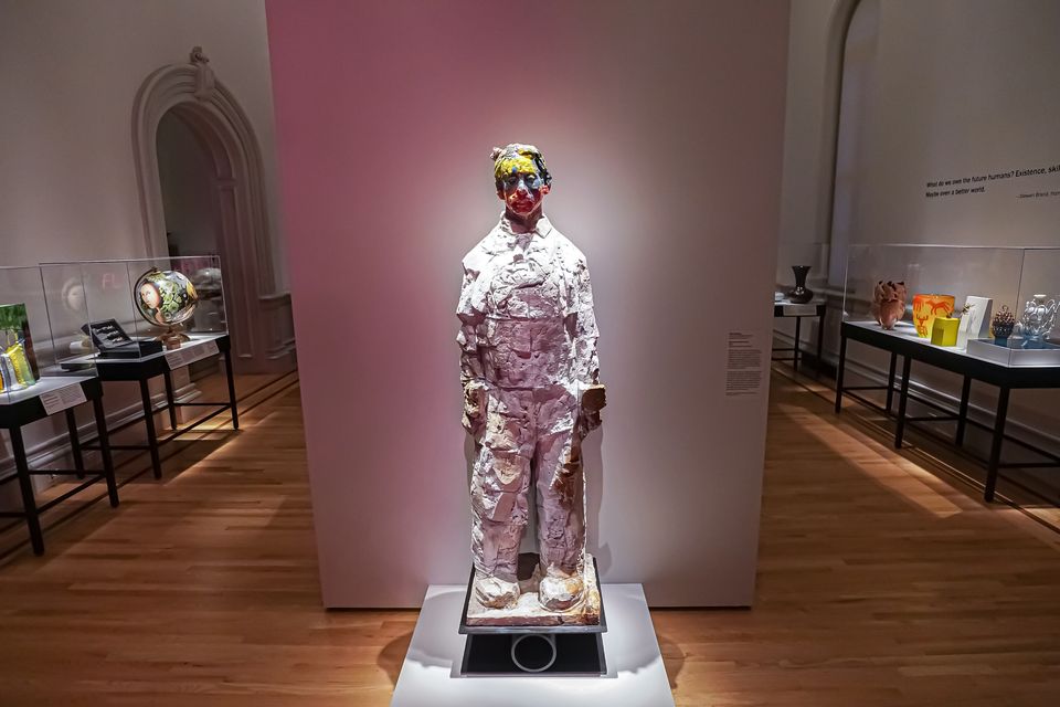 A sculpture in the shape of a person is placed in front of a gallery wall. The face is blurred with different colors.