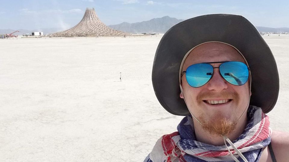 A man wearing a hat, sunglasses, and a scarf taking a selfie in the desert. A pyre is in the distance.