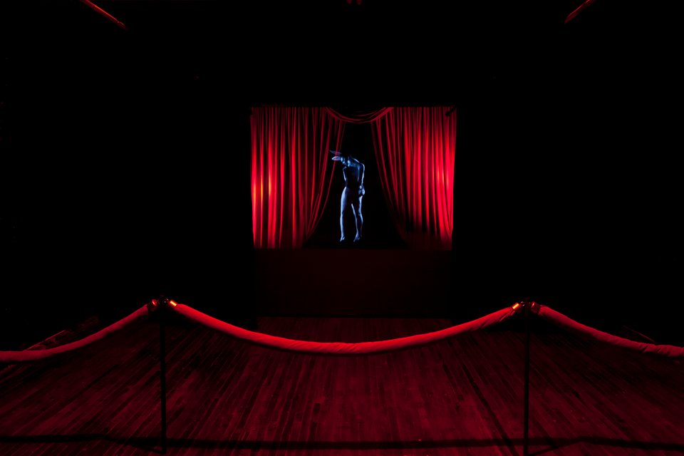 A hollographic figure in a dark room behind a velvet rope and drapes.