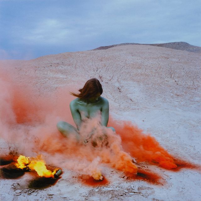 A photograph of a woman sitting down in nature with fire and red smoke around her.