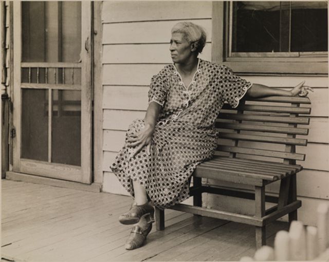 McNeill's gelatin silver print of a woman on a bench outside of a house.