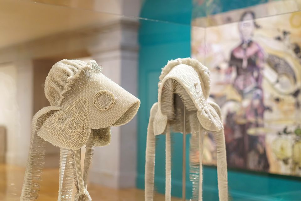 Two off-white, beaded bonnets on display in a glass case. In the background is a large painting.