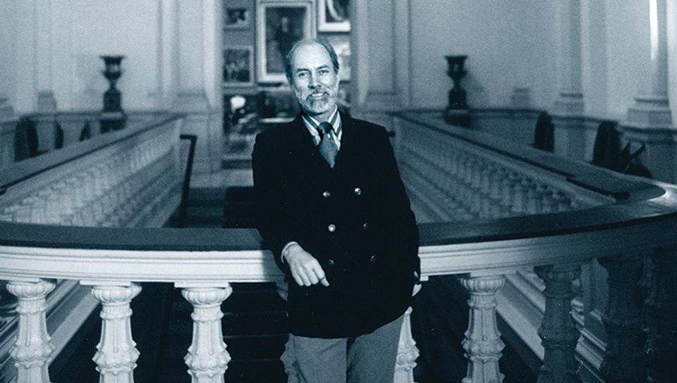 A white man wearing a dark blazer stands leaning on a balustrade. He has a slight smile.