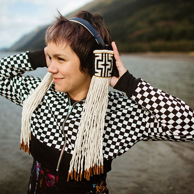 A person in a black and white checkered sweater stands holding headphones. They are also black and white, woven with an Indigenous motif and have long white fringe.