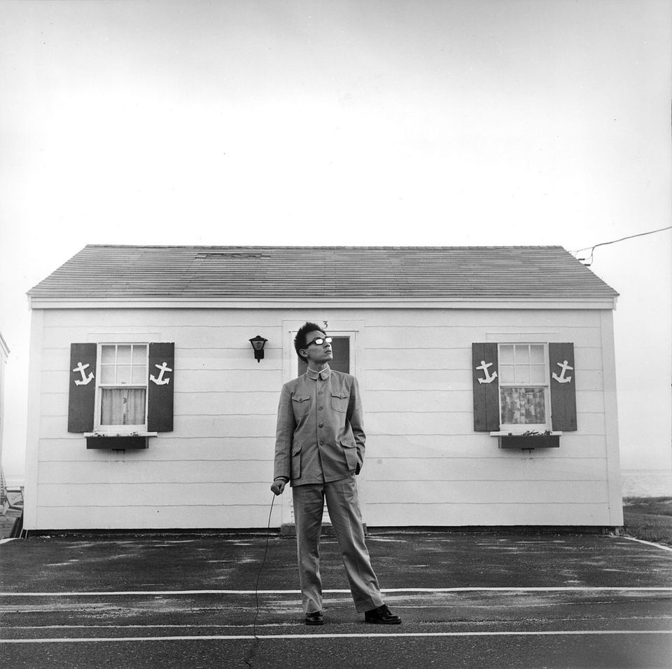 An Asian man with glasses in a suit standing alone in front of a small, white, house.