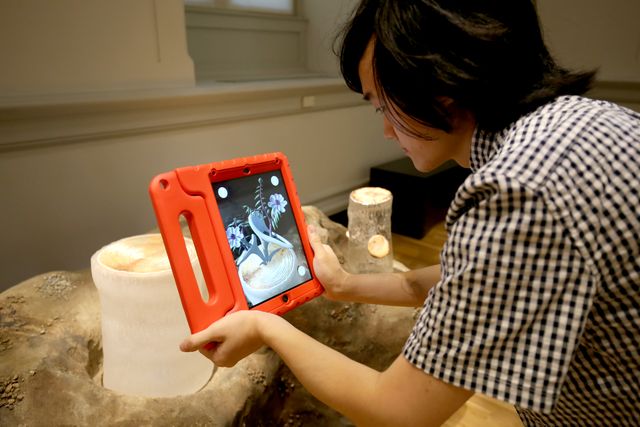 A photograph of a male holding an ipad in front of Ginny Ruffner's artwork.