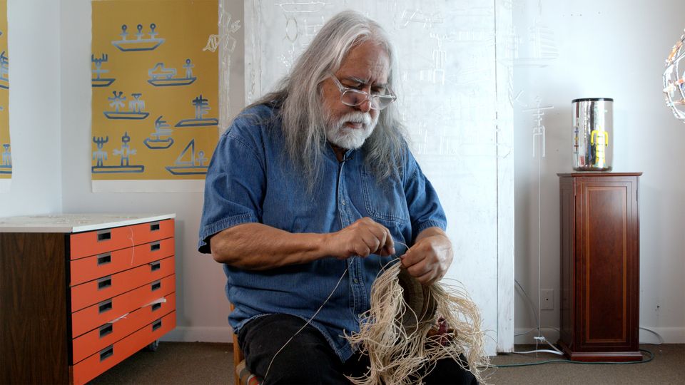 A man with long white hair and a beard sits and works on a straw like material.