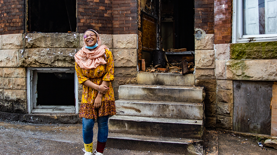 An African American girl wearing a rose-colored hijab stands, smiling, in front of a stone and brick rowhome.