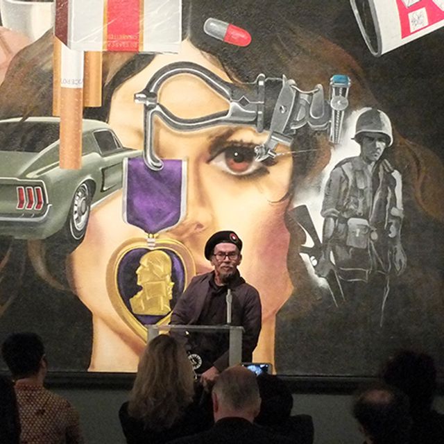 A man wearing a beret stands in front of an artwork. He is speaking to an audience.