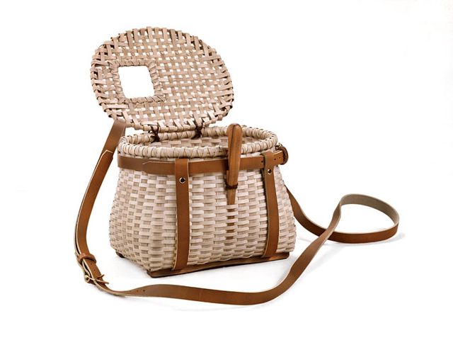A basket with a rectangular base that has a circular top with a lid and leather accents, and a leather strap.