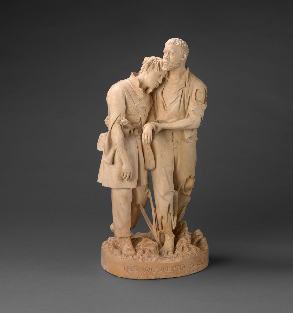 Statue depicting two figures sculpted from a light-colored material against a plain, dark background. One figure is supporting the other who appears injured.  The title on base reads, “The Wounded Scout.”