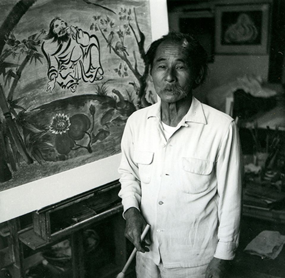 A image of a man in white holding a brush in front of a painting.