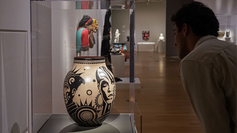 Person leaning toward a vase in a plexiglass covered case in a museum gallery, other artworks fill the space in the distance.