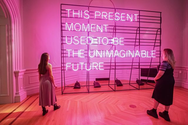 Two people stand in front of a pink neon sign that reads "This Present Moment Used to Be the Unimaginable Future"