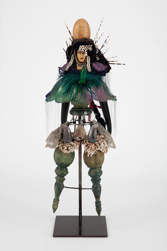 Figure made of assembled objects including cowrie shells, wood, glass horns, rooster feathers, fiber, and more.