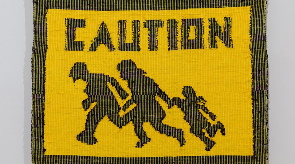 Detail of a yellow tapestry. The word "Caution" is woven into the top and the bottom shows a silhouette of two adults and a child holding hands and running.