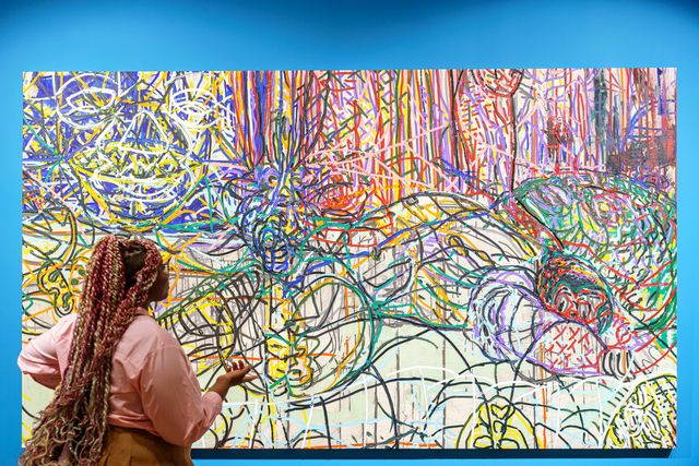 A woman with long braids is looking at a vibrant abstract painting, which hangs on a bright blue wall.