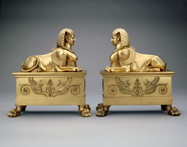 An image of two brass andirons with sphinx tops. 