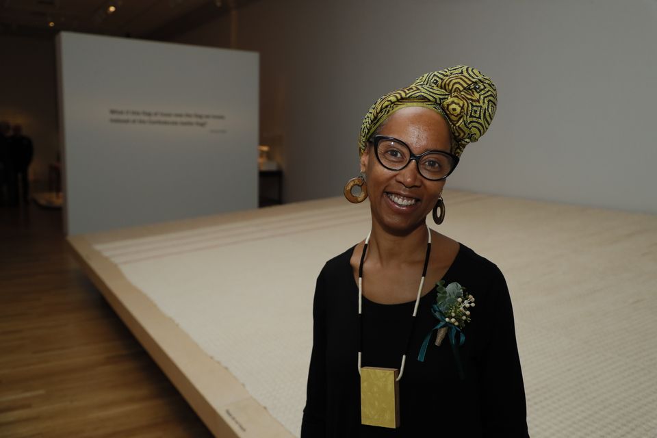An African American woman wearing a headwrap and glasses stands in front of a large-scale woven artwork.