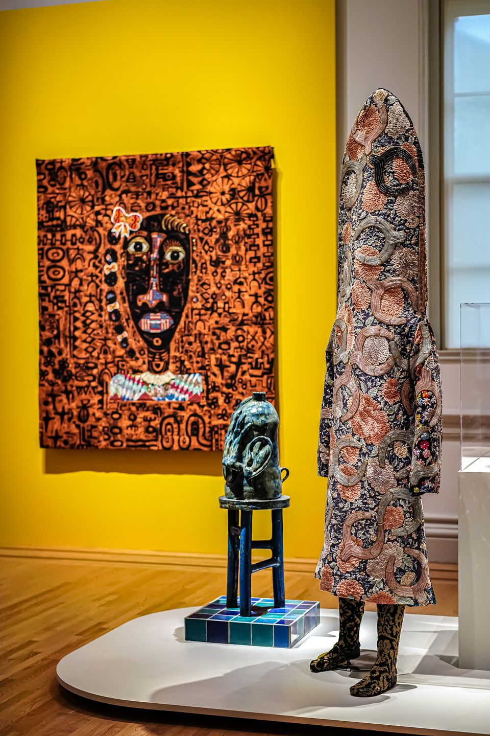 Gallery view of two sculptures standing in front of a quilt, which hangs on a bright yellow wall.