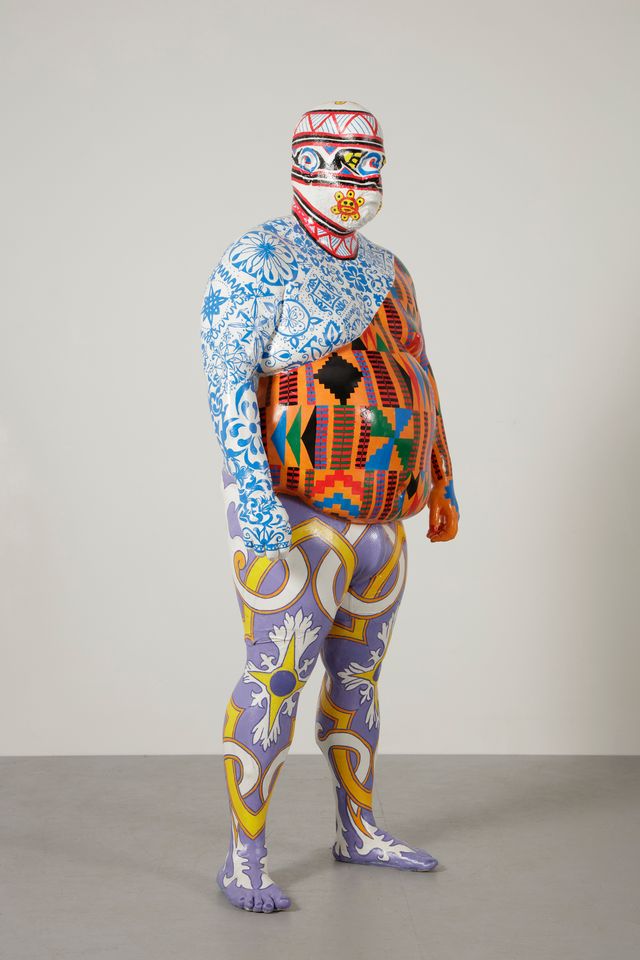 Sculpture of a person completely covered with multiple colorful, intricate patterns standing against a neutral background.