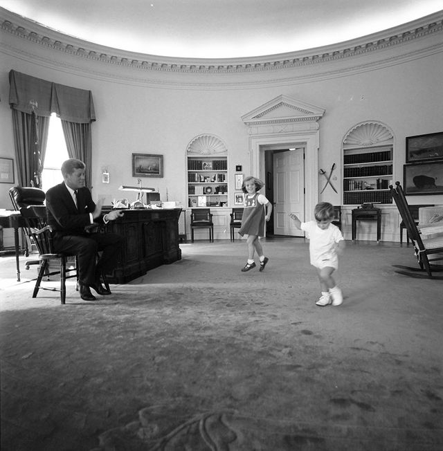 Two small children running across the office while a seated JFK observes them