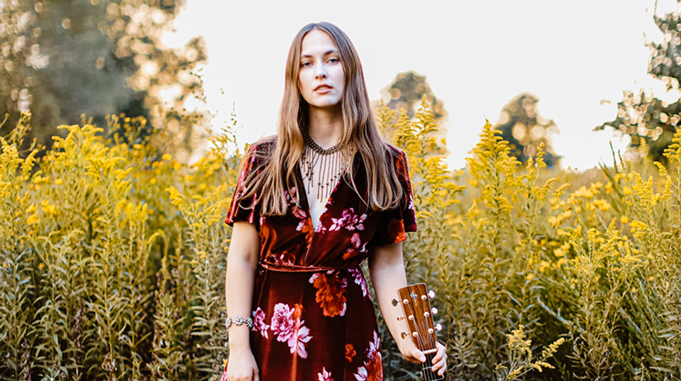 Maddi Mae stands in front of a field of grass wearing a red dress and holding a guitar.