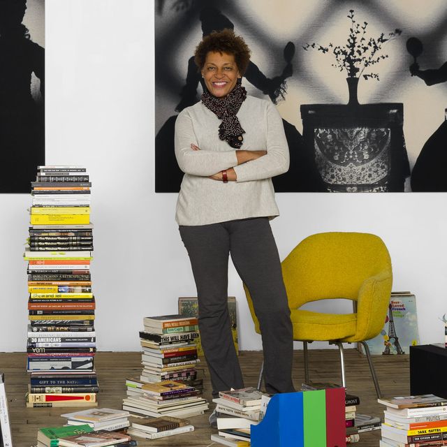 Photo of Carrie Mae Weems standing next to a yellow chair and amidst several stacks of books.