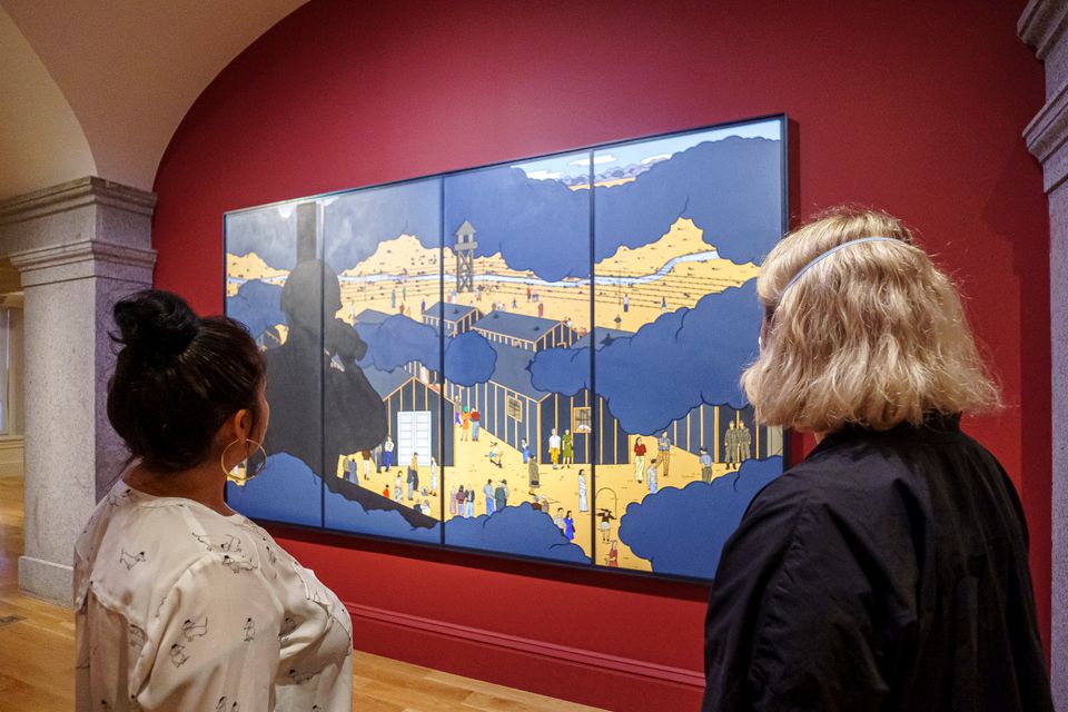 Two people are looking at a painting on a gallery wall. The painting depicts a scene of a work camp and is painted in mostly blues and yellows. It is hanging on a dark red wall.