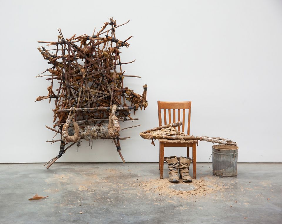 Sculpture formed from sticks, mud, and rope on a wall, with a chair and bucket positioned to the right of it 