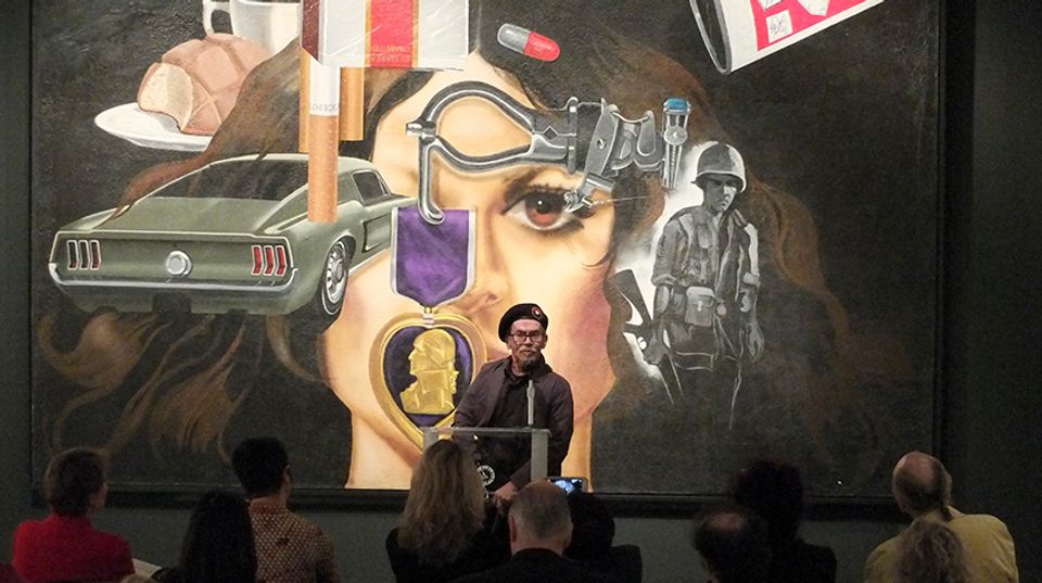 A man wearing a beret stands in front of an artwork. He is speaking to an audience.
