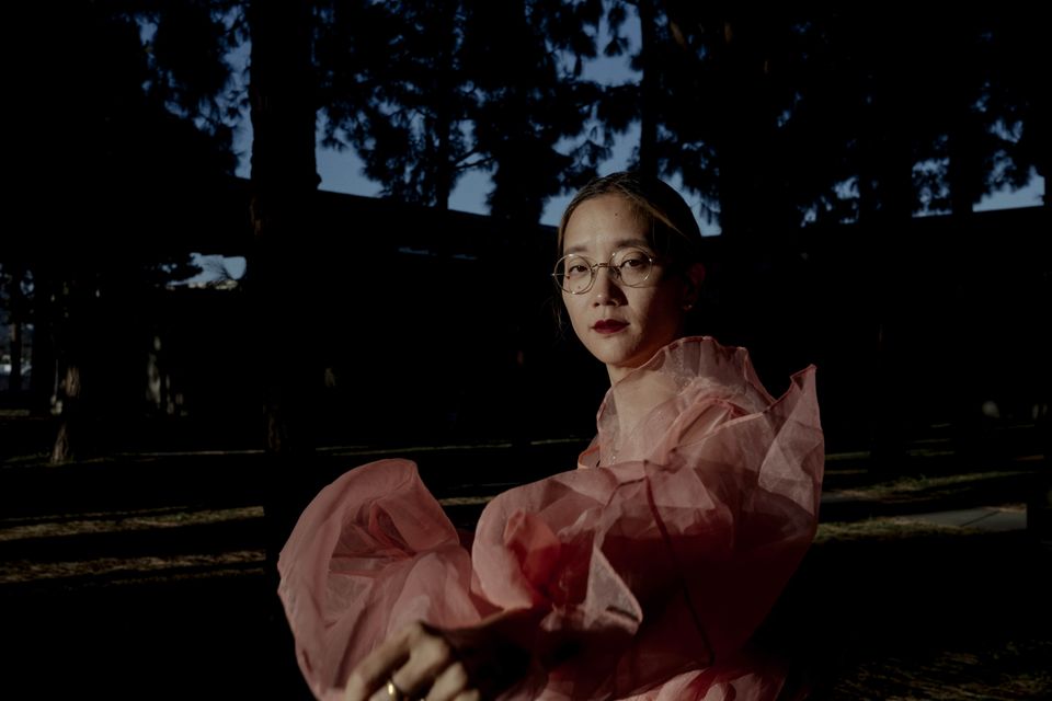 Christine Sun Kim stands in front of a tree and dark background in a pink dress