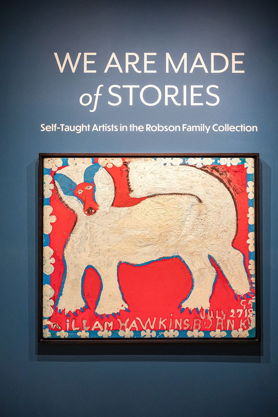 Blue wall with a painting of a dog hanging and the title above it reads "We Are Made of Stories: Self-Taught Artists in the Robson Family Collection."