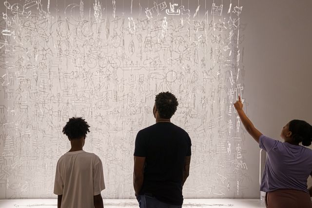 Three young people stand in front of an art installation made of hanging clear glass charms. The figure on the right is point up at the artwork.