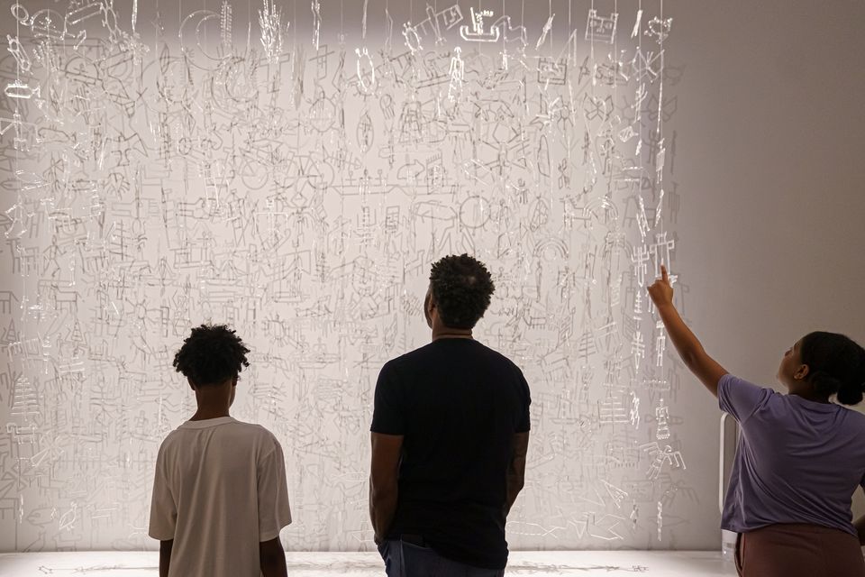 Three young people stand in front of an art installation made of hanging clear glass charms. The figure on the right is point up at the artwork.