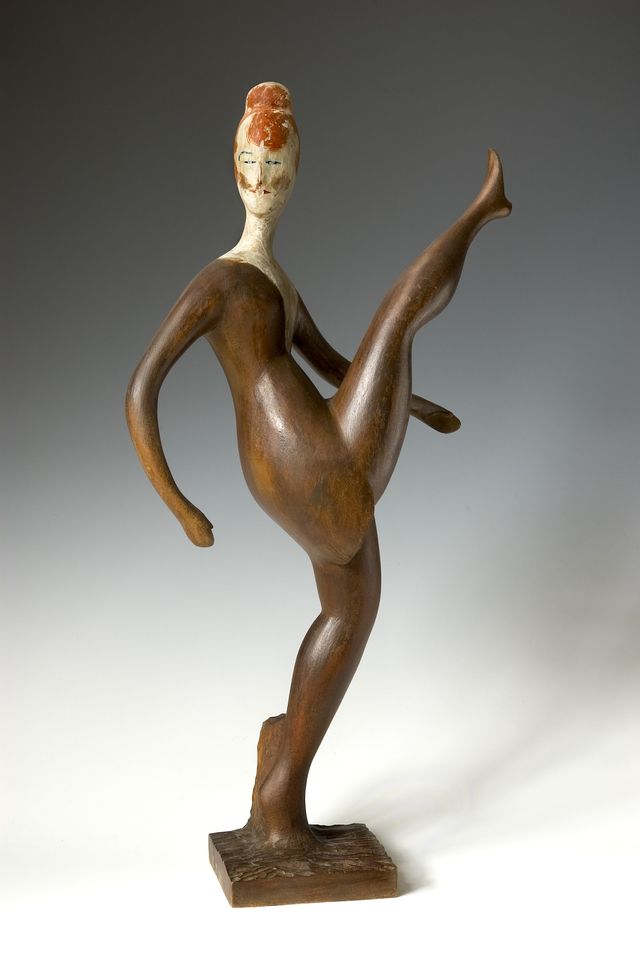 A sculpture made of wood of a dancer kicking their leg up in the air.
