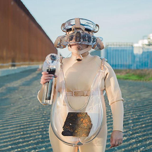 Artist Tanya Aguiñiga wearing a suit made of glass elements while standing next to the U.S./Mexico border wall