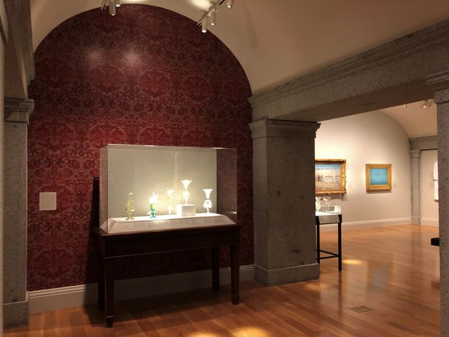 Gallery image showing a case of glass artworks in front of a dark red wall.