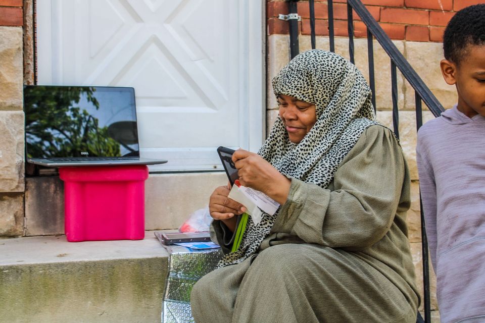 An African American woman wearing a leopard print hijab looks at her phone and smiles. Resting on the top step to her right is a laptop. A boy stands half inside the frame to her left.
