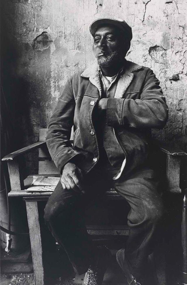 A gelatin silver print of a man sitting and smoking with one hand in his jacket.