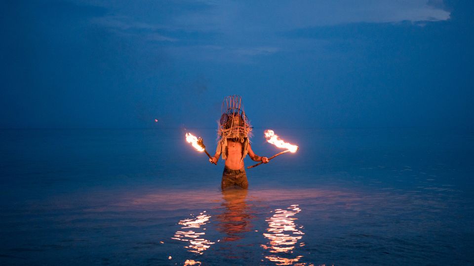 A person stands in the ocean. He is wearing a headdress and is holding a flaming torch in each hand.