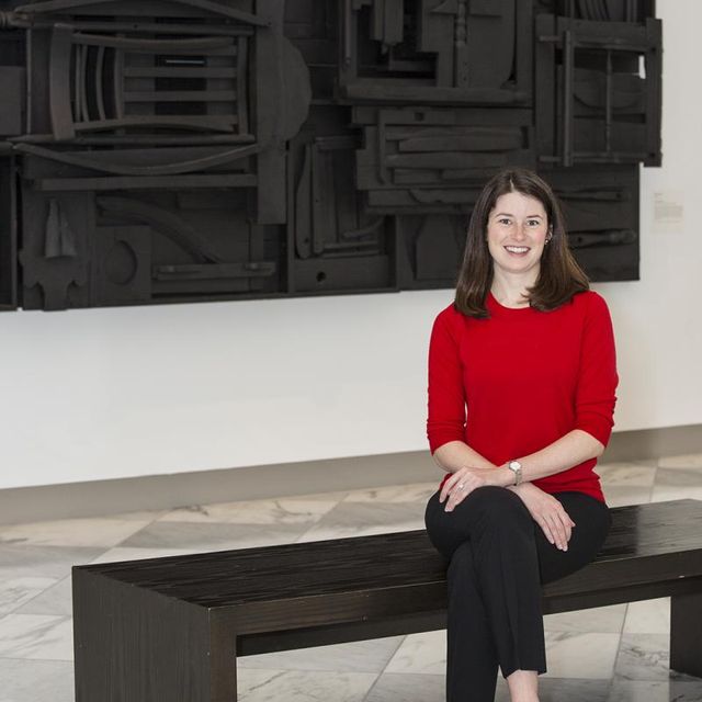 A photograph of a woman sitting down on a bench in a gallery with a red sweater on and a black artwork behind her.