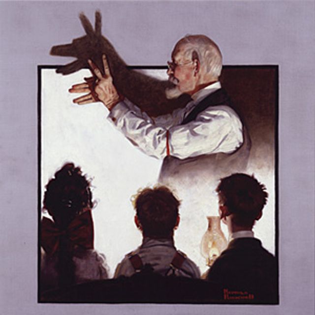 An image of a man making shadow puppets on the wall in the background and three children watching in the foreground. 